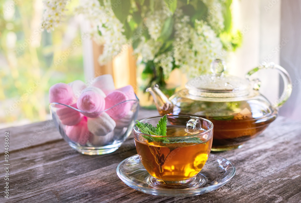 Mint tea in glass Cup on the background of a vase with marshmallows and a transparent glass teapot in a Sunny summer morning. Mint tea in glass Cup on the background of a vase with marshmallows and a transparent glass teapot in a Sunny summer morning.