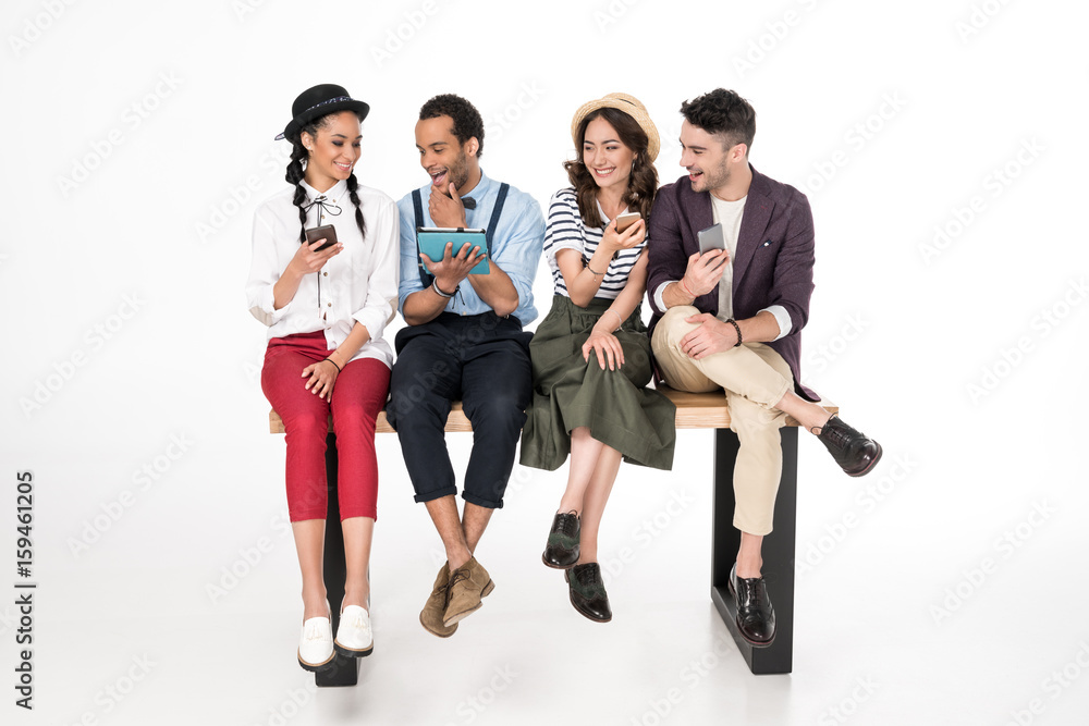 Smiling young men and women sitting together on bench and using digital devices Smiling young men and women sitting together on bench and using digital devices