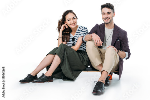 smiling couple sitting on skateboard and looking at camera isolated on white