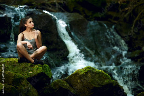 Hiking in forest with waterfall in background. Woman trekking in mountains against waterfall. Female with backpack on hike in nature. Cascading waterfall