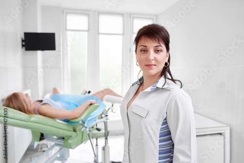 Confident female doctor posing at her clinic female patient lying on gynecological chair on the background waiting for medical examination people job occupation health medicine concept.