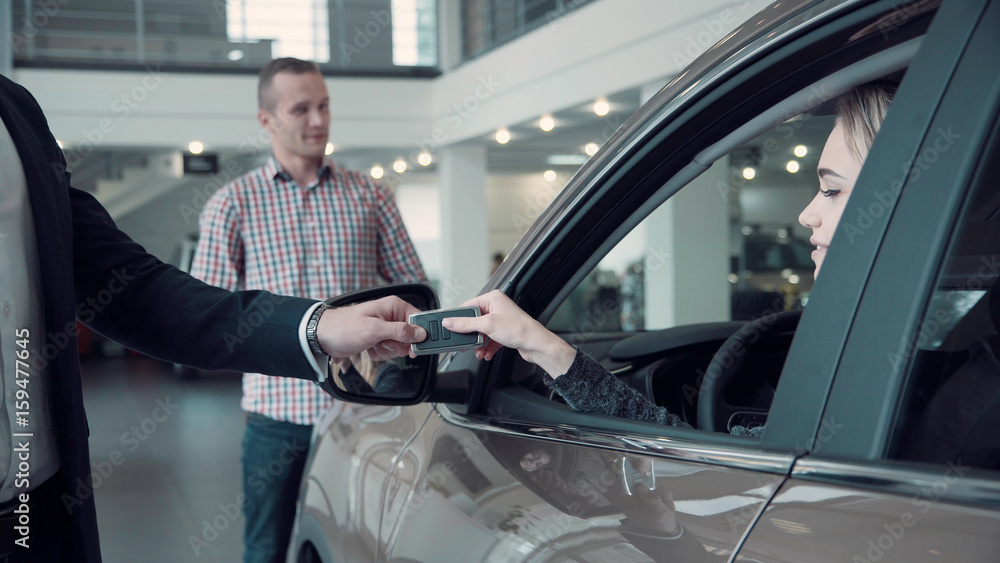 Unrecognizable Sales Manager Gives the Client the Keys from the Car. Then smiling woman customer look straight at the camera, while her husband standing in the background