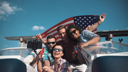 Emotional group of mixed ethnic young friends on board a motor boat along a river laughing and cheering and raise american flag in air. Celebrate an Independence Day on July 4