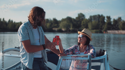 Schilderij op canvas Young couple toasting each other with cans of beer as they relax in the evening on a lake in a motorboat while enjoying their summer vacation