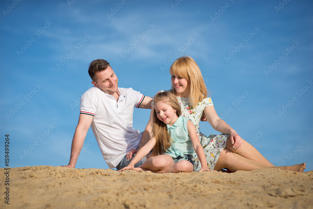 Happy beautiful laughing family sitting on the sand on the beach against the background blue sky in summer vacation. Happy beautiful laughing family sitting on the sand on the beach against the background blue sky in summer vacation.
