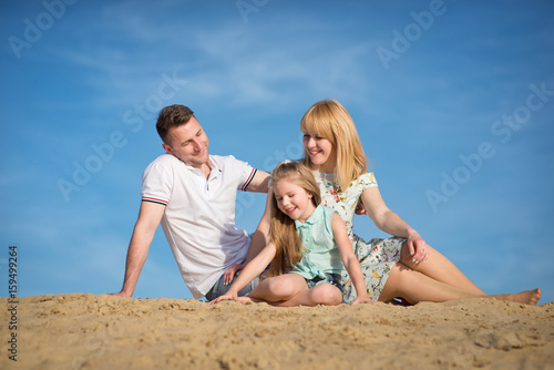 Happy beautiful laughing family sitting on the sand on the beach against the background blue sky in summer vacation.