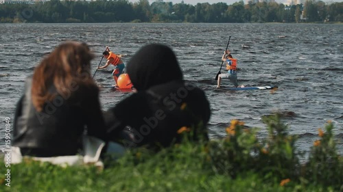 Couple sitting on a grass watch as two surfers rides across water in front of them on a summer windy day