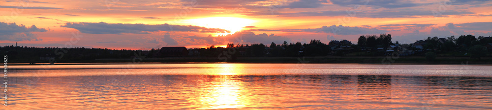 Fototapeta premium Panorama zachodu słońca z widokiem na jezioro i dom oraz drzewa na przeciwległym wybrzeżu.