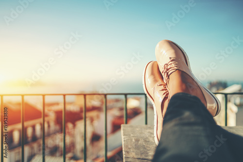Female feet with glossy pale pink shoes relaxing on wooden table and touristic european city near sea with multiple houses in blurred background with copy space for advertising, logo or your texts photo