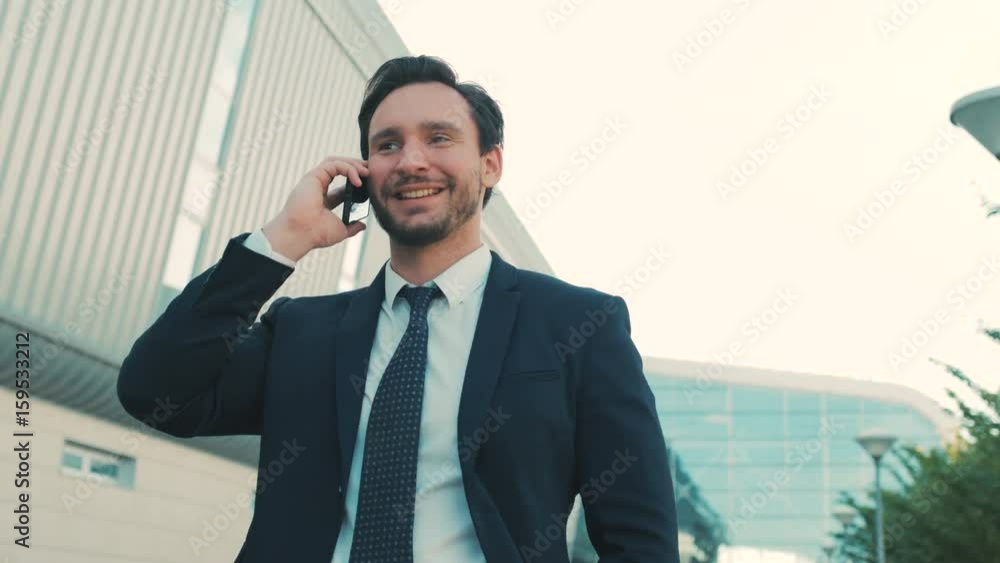 Handsome businessman talking on the cell phone while walking outdoors near office center and looking straight at the camera and smiling. Portrait shot, close up Handsome businessman talking on the cell phone while walking outdoors near office center and looking straight at the camera and smiling. Portrait shot, close up