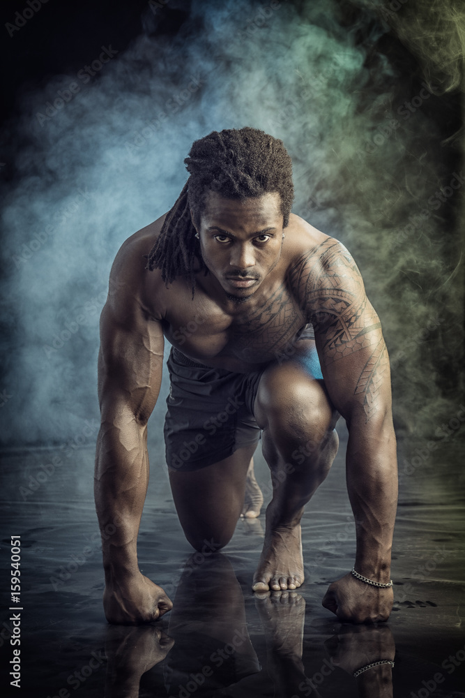 Handsome young muscular black man shirtless ready to sprint and run. On dark background with smoke Handsome young muscular black man shirtless ready to sprint and run. On dark background with smoke