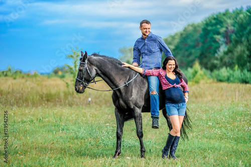 Awaiting the child, walking on the meadow. Young couple - she is a handsome brunette with long hair, pregnant  he is tall and brave, astride a black horse. © schamin