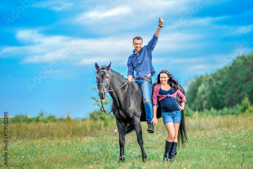 Awaiting the child, walking on the meadow. Young couple - she is a handsome brunette with long hair, pregnant  he is tall and brave, astride a black horse. © schamin