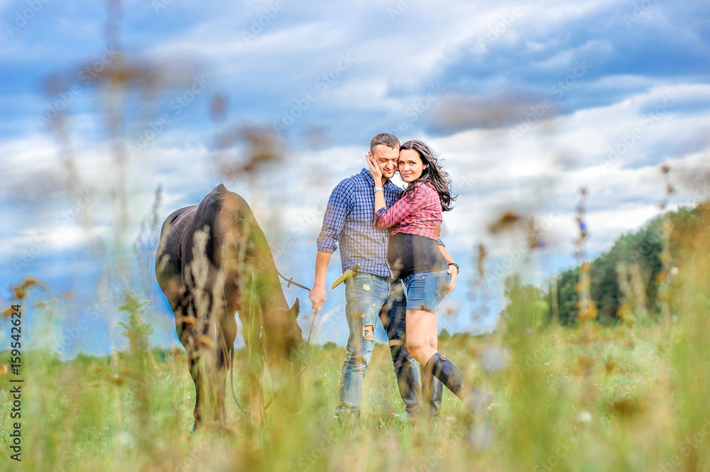 View through the grass: young couple - she is handsome brunette with long hair, pregnant; he is tall and brave, holding the reins of the black horse, a walk on the meadow. Wonderful sky behind them