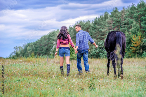 Awaiting the child. Young couple - she is a brunette with long hair, pregnant; he is tall and brave, holding the reins of the black horse, a walk on the ranch.