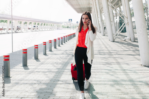 Woman with red suitcase walks along the airport and talks on the phone