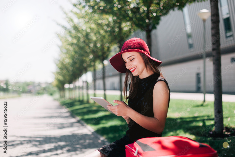 Woman in red hat checks her tablet sitting by a red suitcase Woman in red hat checks her tablet sitting by a red suitcase