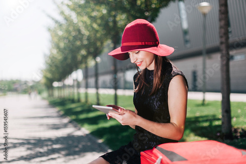 Woman in red hat checks her tablet sitting by a red suitcase