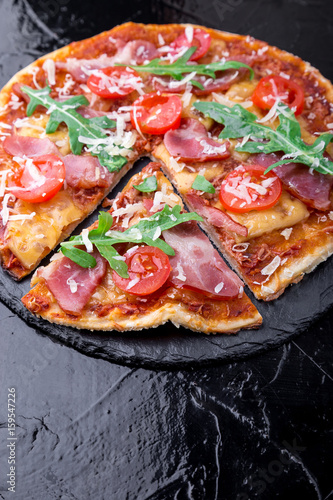 Homemade pizza with prosciutto, tomato, arugula on black slate board. Close up.