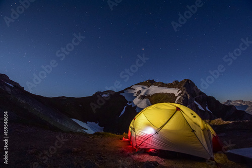 Illuminated tent on a rock near the summit of Westliche Karwendelspitze. Moonlit mountain in the background. Stars in sky. Karwendel is a mountain range in Bavaria, Germany and Tirol, Austria.