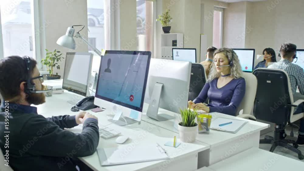 PAN of male and female call centre operators in headsets sitting before computers in airy office and talking to clients via online voice calls PAN of male and female call centre operators in headsets sitting before computers in airy office and talking to clients via online voice calls