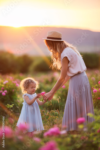 Beautiful child girl with young mother are wearing casual clothes walking in roses garden over sunset lights