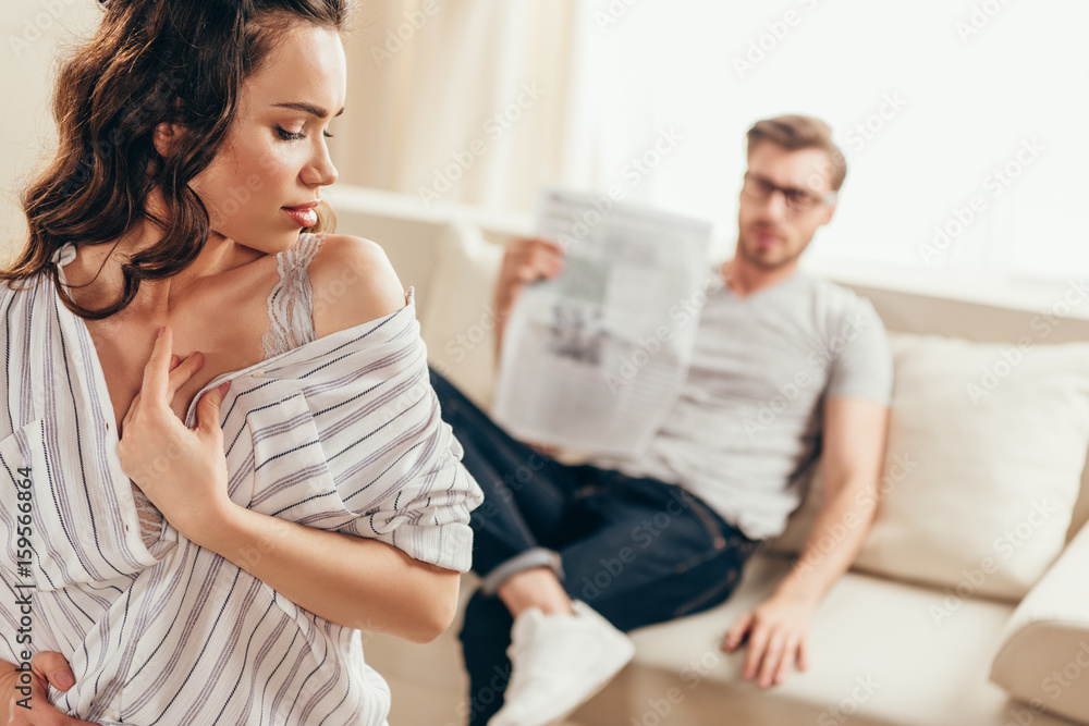 Close-up view of beautiful sensual young woman flirting with boyfriend sitting on sofa and reading newspaper