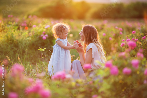 Beautiful child girl with young happy mother are wearing casual clothes walking in roses garden over sunset lights, summer time