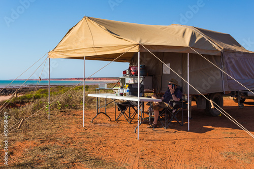 Man using mobile telephone under awning of an off road camper trailer set up on the cliffs at James Price Point.