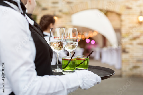 Midsection of professional waiters in uniform serving wine, cocktails and snacks during buffet catering party, festive event or wedding. Full glasses of wine on tray. Outdoor party catering service.