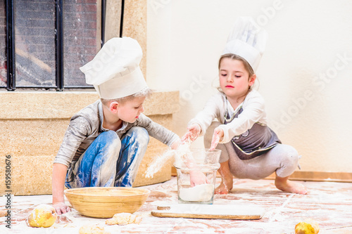 Two siblings - boy and girl - in chef's hats near the fireplace sitting on the kitchen floor soiled with flour, playing with food, making mess and having fun