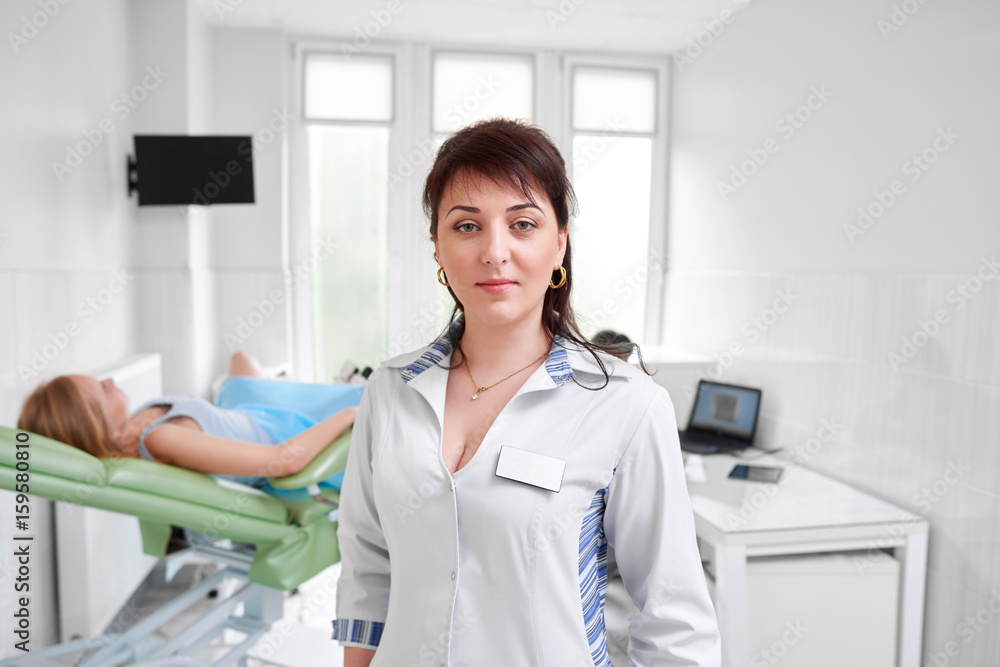 Mature female doctor posing confidently at the hospital female patient lying on gynecological chair on the background medicine healthcare feminine health gynecology professionalism trust.