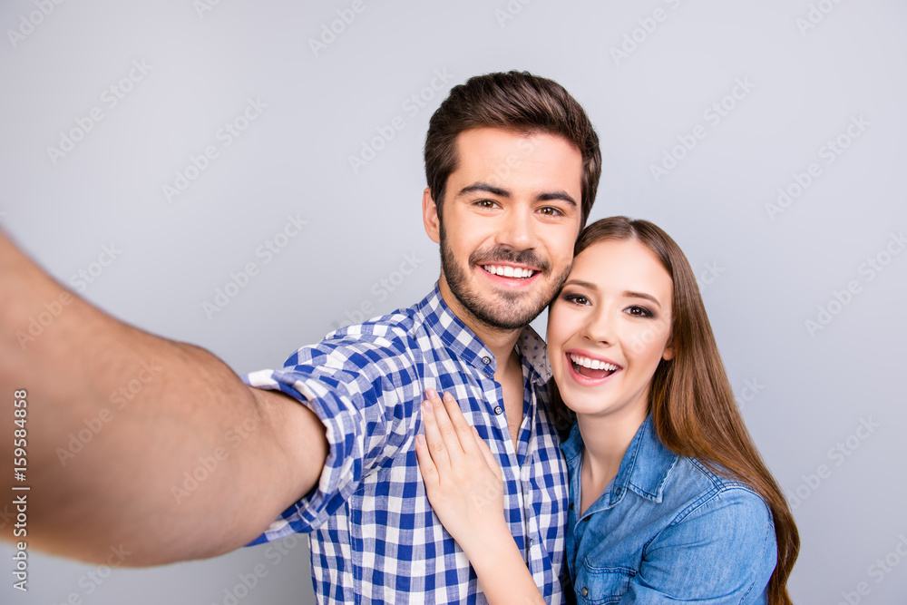 Close up of a cheerful young couple making selfie photo on guy`s camera. They are in casual outfits, posing and smiling on pure light background, embracing Close up of a cheerful young couple making selfie photo on guy`s camera. They are in casual outfits, posing and smiling on pure light background, embracing