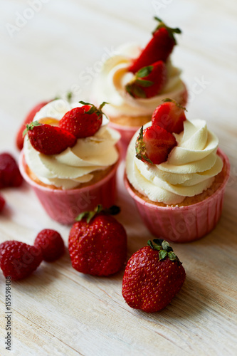 Cupcakes with whipped cream and sweet cream, decorated with strawberries.On a light wooden background.