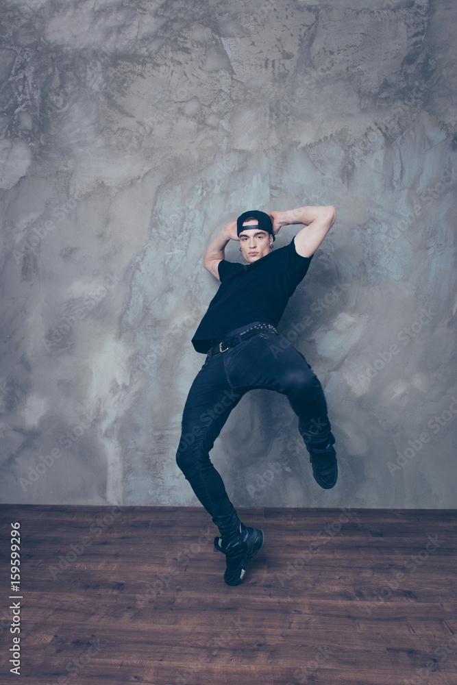 Portrait of a dancing young man in black outfit and cap on grey background