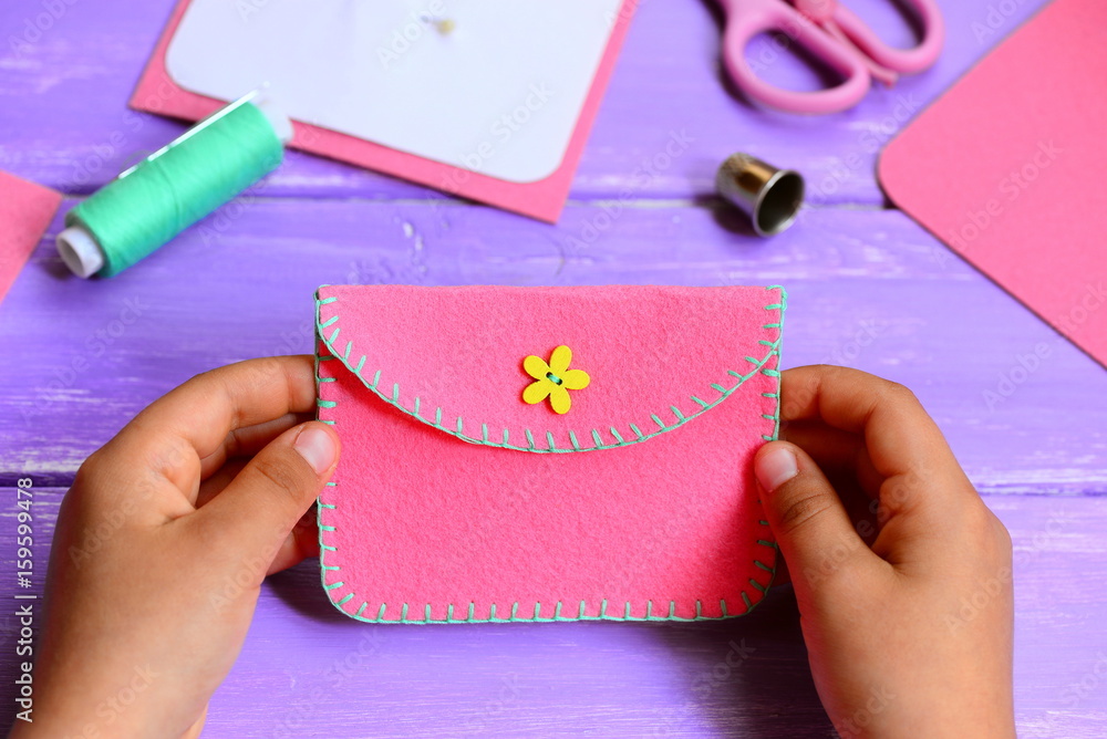 Obraz premium Child made a felt purse. Small child holds a pink felt purse in his hands. Craft supplies on a wooden table. Fun and easy sewing project for kids