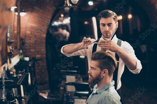 Profile view of a red bearded stylish barber shop client. He is getting his perfect trendy haircut from a classy dressed handsome stylist, looking in a mirror and waiting for result