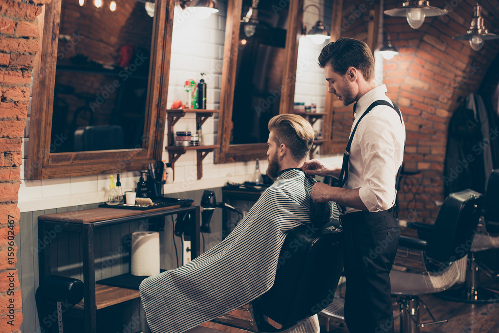 Back view of  a hairstylist and a client in a barber shop. Bearded young man with stunning hairdo is going to have his beard styling. Hairdresser is putting the cape on him