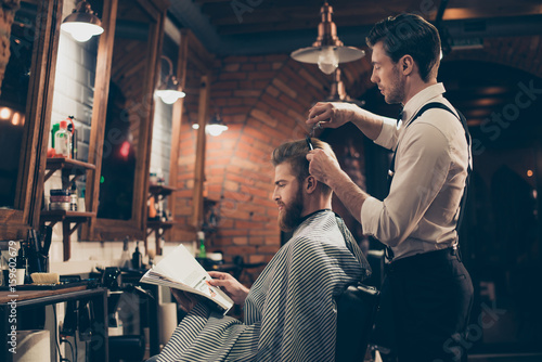 Low angle profile view of red bearded stylish barber shop client, which is getting his perfect haircut from a classy dressed stylist, reading the magazine and waiting for result