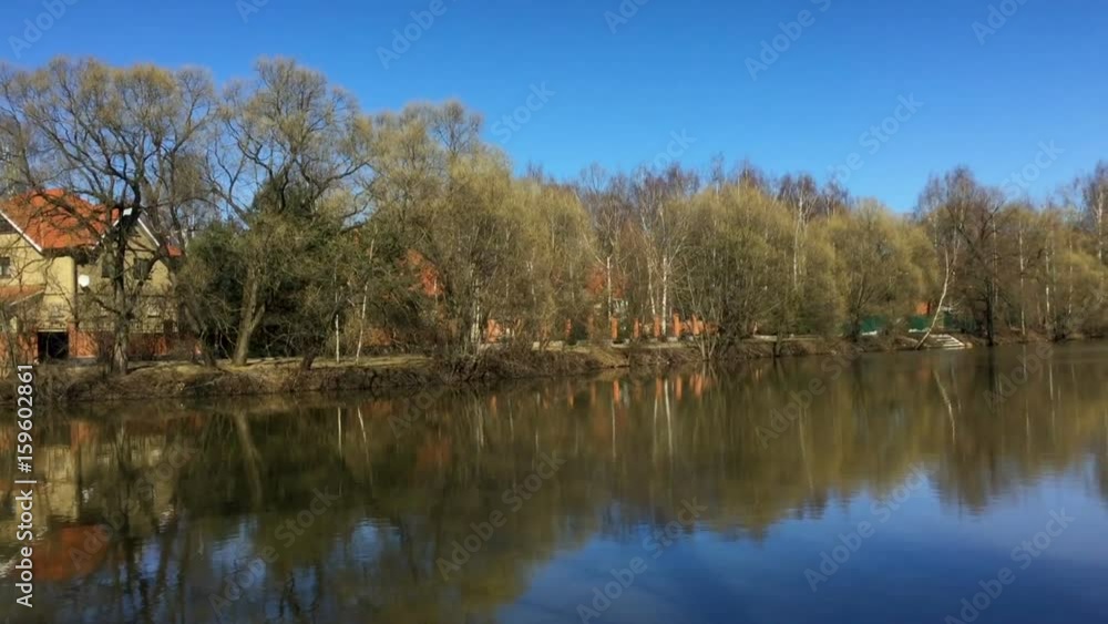 Serene early spring landscape of countryside with pretty house on riverside and water reflecting bright blue sky and bare trees Serene early spring landscape of countryside with pretty house on riverside and water reflecting bright blue sky and bare trees