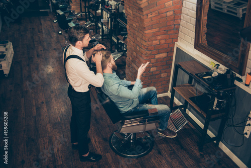 Top view of barber shop classy dressed stylist, who is working for a perfect hairdo of a blond bearded guy in caual jeans outfit. Both looking in the mirror, client is explaining what he wants