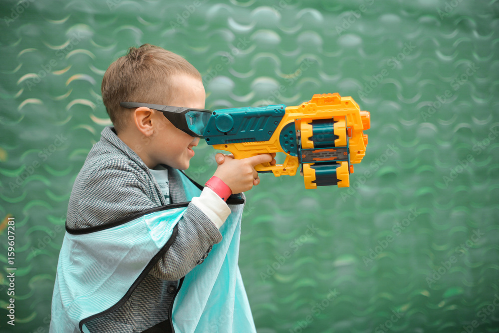 Naklejka premium Small preschooler boy with blaster prepare for attack and play with friends in protective glasses. Excited Child with darts toy gun on the play field