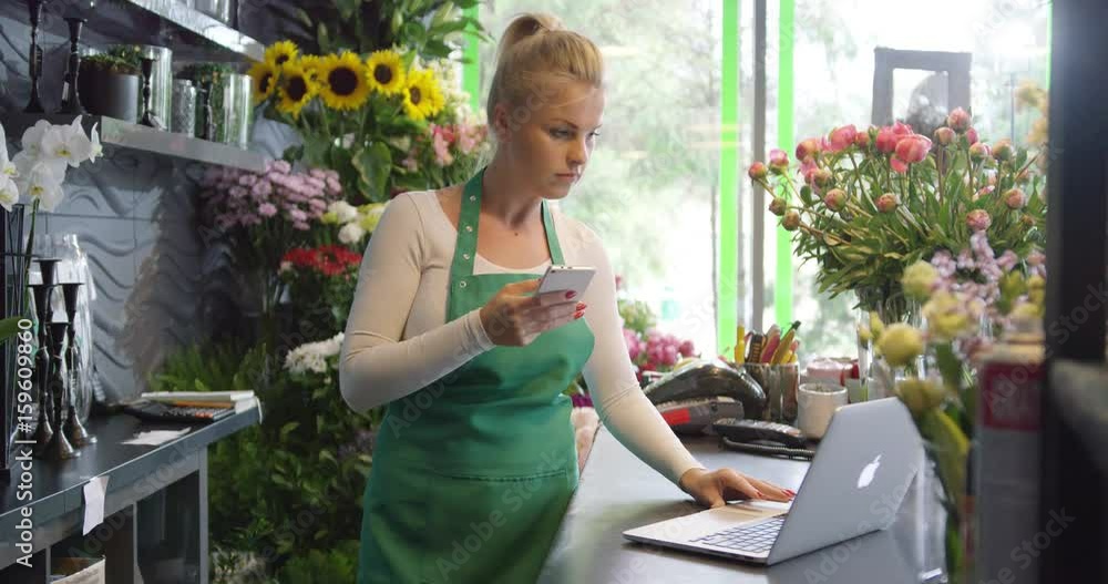 Side view of young woman in green apron standing with smartphone in hands behind counter and browsing laptop. Side view of young woman in green apron standing with smartphone in hands behind counter and browsing laptop.