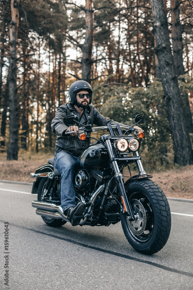 Biker man wearing jeans and leather jacket riding on motorcycle. Forest freedom scene. Biker man wearing jeans and leather jacket riding on motorcycle. Forest freedom scene.