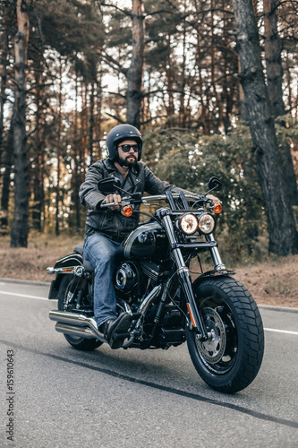 Biker man wearing jeans and leather jacket riding on motorcycle. Forest freedom scene.