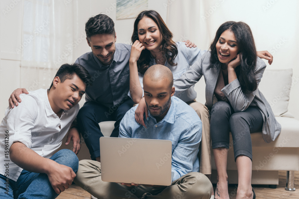 Fototapeta premium young smiling multiethnic friends looking at laptop while sitting on sofa at home