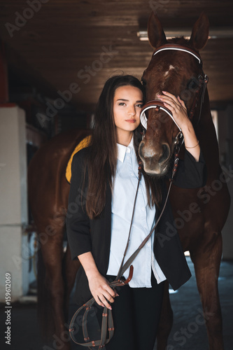 Pretty young woman standing in barn hugging her horse