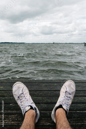 Man sitting on pier with sneakers on foreground and seascape on background