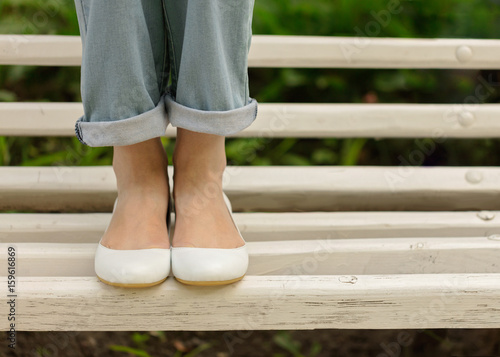 Slika na platnu Female legs in blue jeans and white shoes on a white bench.