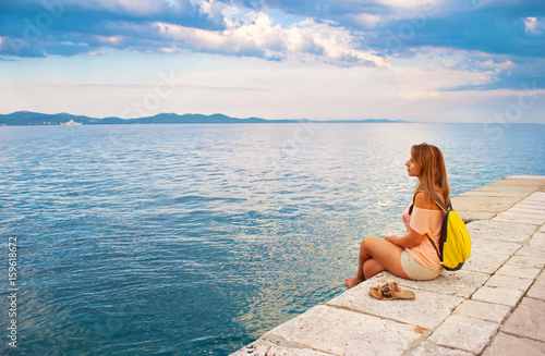 One young girl with a backpack sitting peacefully sideways near the sea shore looking wistfully at the hill range in the distance, her legs crossed. Zadar, Croatia